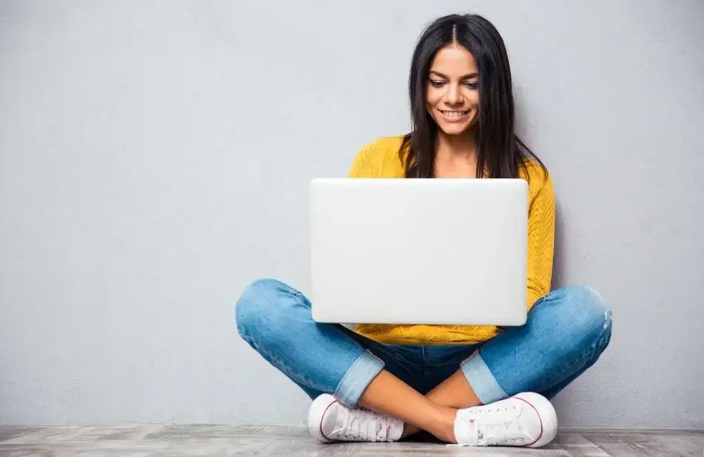 Happy young woman sitting on the floor with crossed legs and using laptop on gray background